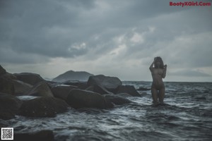 A woman in a white tank top sitting on a rock by the ocean.
