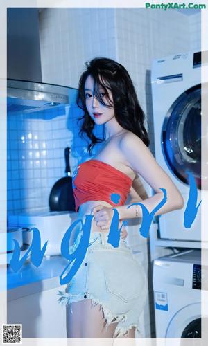 A woman in a red top and denim shorts standing in front of a washing machine.